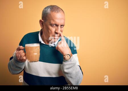 Senior handsome man drinking jar of beer standing over isolated yellow background Pointing to the eye watching you gesture, suspicious expression Stock Photo