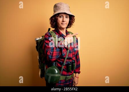 Middle age curly hair hiker woman hiking wearing backpack and water canteen using binoculars Pointing with hand finger to the side showing advertiseme Stock Photo