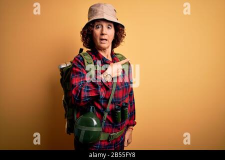 Middle age curly hair hiker woman hiking wearing backpack and water canteen using binoculars Surprised pointing with finger to the side, open mouth am Stock Photo