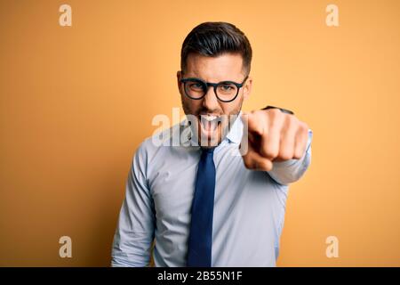 Young handsome businessman wearing tie and glasses standing over yellow background pointing displeased and frustrated to the camera, angry and furious Stock Photo