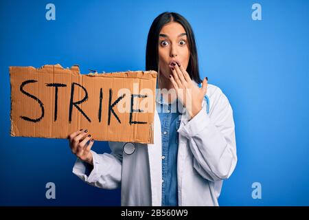 Young doctor woman wearing stethoscope holding cardboard banner protesting in strike cover mouth with hand shocked with shame for mistake, expression Stock Photo
