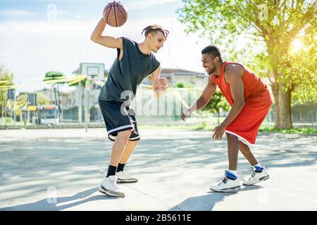 Two multiracial cheerful friends playing basketball outdoor in city urban contest with back light - Young people having fun doing sport for a healthy Stock Photo