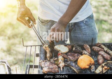 Afro american man cooking meat on barbecue - Chef putting some sausages on grill in park outdoor - Concept of eating outdoor during summer time - Vint Stock Photo