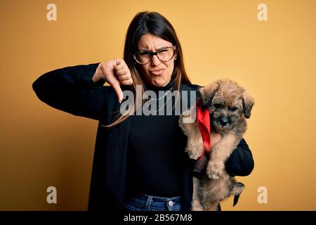 Young beautiful brunette woman holding cute puppy pet over isolated yellow background with angry face, negative sign showing dislike with thumbs down, Stock Photo