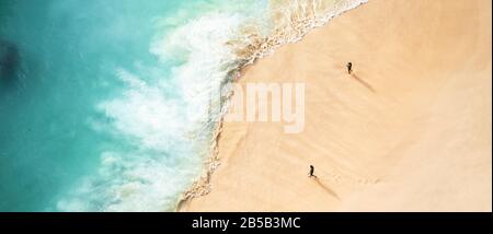 View from above, stunning aerial view of two people walking on a beautiful beach bathed by a turquoise sea during sunset. Stock Photo