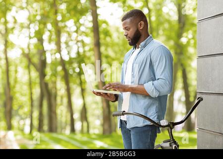 Handsome Black Guy Standing With Laptop Outdoors In Park, Checking Wifi Stock Photo