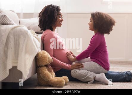 Little afro girl touching her pregnant mom belly, feeling baby kicks Stock Photo