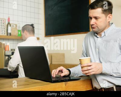 Serious millennial man checking data at laptop and reads in the coffeeshop. Hipster in the cafe reading news. Stock Photo