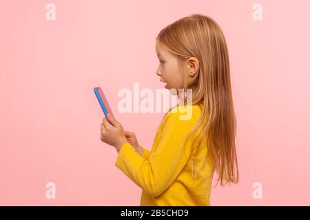 Wow, great app for kids. Side view of astonished little girl using smartphone with shocked expression, playing game on mobile device, pleasantly surpr Stock Photo