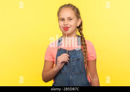 Portrait of charming happy little girl with braid in denim overalls covering mouth with red paper lips on stick and smiling at camera, symbol of kiss. Stock Photo