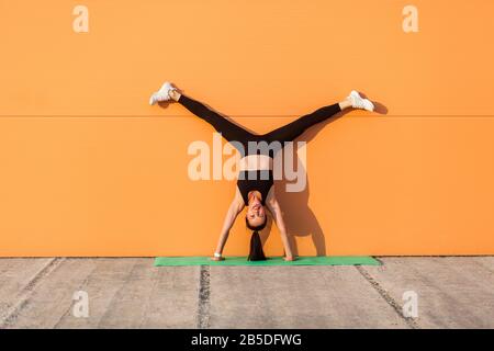 Overjoyed happy girl with perfect athletic body in tight sportswear doing yoga handstand pose with spread legs against wall and showing tongue, having Stock Photo