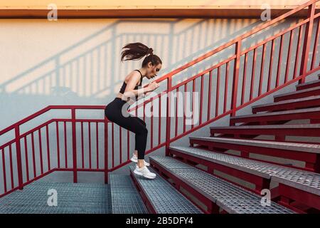 Fit athlete beautiful woman in tight sportswear jumping on stairs, warming up before jogging, motivated young person full of energy training on stairc Stock Photo