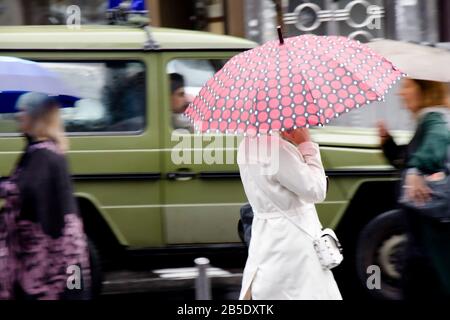 Belgrade, Serbia - September 24, 2019 : Young  woman in white rain coat walking under umbrella on a rainy city street on a busy day in motion blur Stock Photo