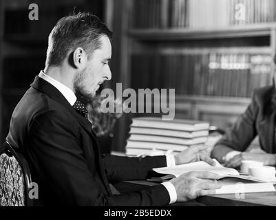 Science and studying concept. Man in classic suit or professor with concentrated face sits in library near piles of books, bookshelves on background, reading book, defocused. Men study in old library. Stock Photo