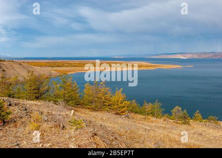 Autumn landscape overlooking Lake Baikal. Yellow trees and blue water. Stones on the ground. Clouds in the sky and rain in the distance. Horizontal. Stock Photo