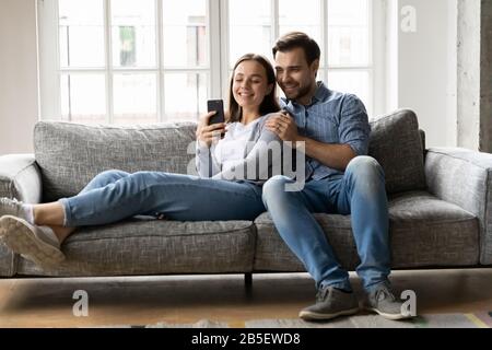 Happy young couple relaxing on sofa, looking at cellphone screen. Stock Photo