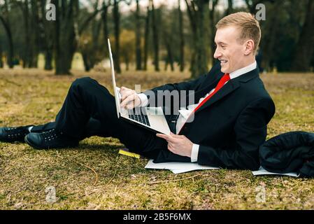 young guy lying on the nature with a laptop. businessman working remotely outdoors. freelancer communicates over the network Stock Photo