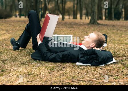 young guy lying in nature with laptop. businessman working remotely outdoors. freelancer reads documents. Stock Photo