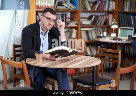 Man reading book and drinking coffee in a library cafe with many books on a background Stock Photo