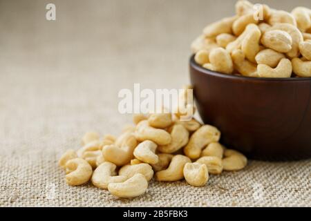 Cashew nuts in a wooden bowl on a burlap cloth background. Golden cashew close-up in a dark brown cup. Stock Photo