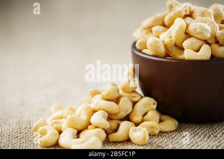 Cashew nuts in a wooden bowl on a burlap cloth background. Golden cashew close-up in a dark brown cup. Stock Photo