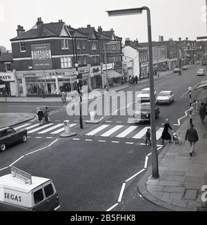 Aerial view of a new road safety measure, a zebra or pedestrian crossing on a busy south london high street, circa late 1960s. First introduced in the UK in 1949, a level crossing gives priority to pedestrians and were put in places where there were shops, schools and other amenities used by pedestrians. Motorists have to give way.  Motorcars of the era can be seen, with a Segas van on the left. Stock Photo