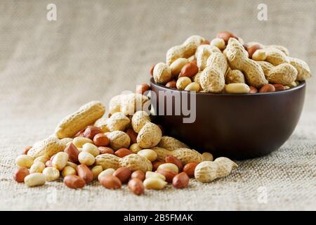 Peanuts in the shell and peeled close up in a cup. Background with peanuts. Roasted peanuts in the shell and peeled on a background of brown cloth in Stock Photo