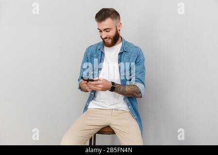 Portrait of a handsome smiling young man wearing casual clothes sitting isolated over gray background, using mobile phone Stock Photo