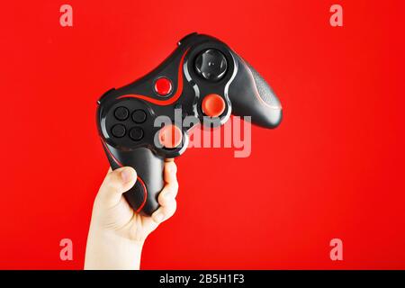 A child's hand triumphantly holds the gamepad on a red background. Minimalistic creative concept of the younger generation of gamers. Winning online v Stock Photo