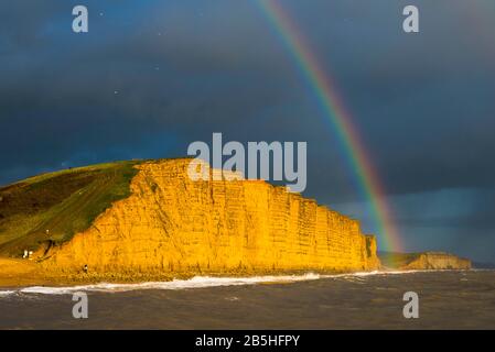 West Bay, Dorset, UK.  8th March 2020.  UK Weather.  A rainbow arches above the cliffs which are illuminated by late afternoon sunshine at West Bay in Dorset as a dark shower cloud passes over.  Picture Credit: Graham Hunt/Alamy Live News Stock Photo