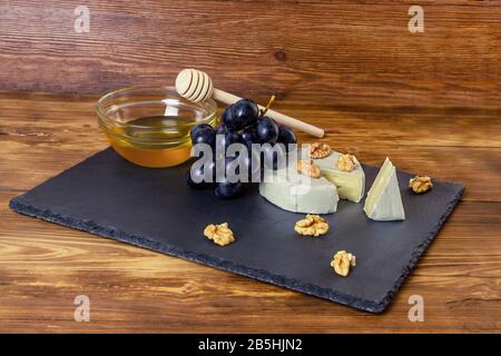 Camembert cheese in a dark tray next to sweet blue grapes nuts and honey on wooden background Stock Photo