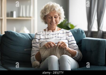 Happy 60s grandmother relax on couch doing knitting Stock Photo