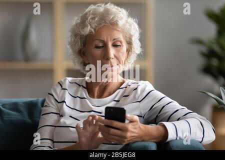 Modern elderly woman sit on couch texting on cell Stock Photo