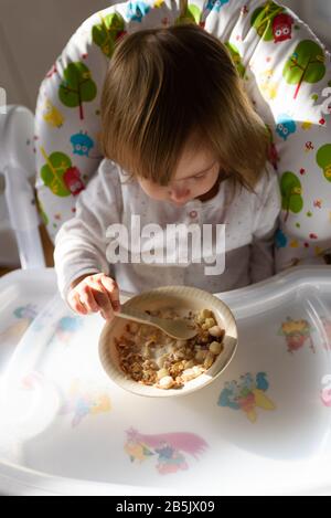 Two years old eats brakefast by herself with a spoon. Stock Photo