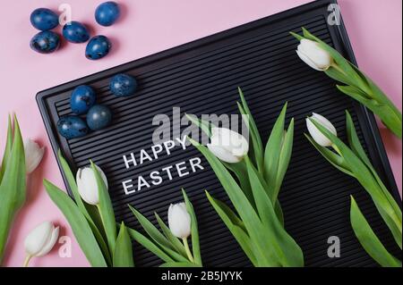 Happy Easter - text on black letter board with white tulips and blue colored eggs on pink background. Stock Photo