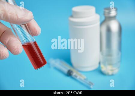 A hand in gloves holds a test tube with red tests on the background of medical cans, pills on a blue background. close up. Stock Photo