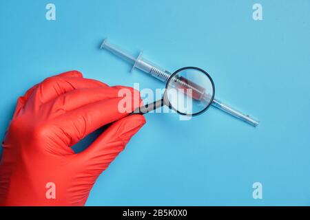 Hand in gloves holds a magnifying glass over a syringe with a vaccine. Drug research concept. Close up. Stock Photo