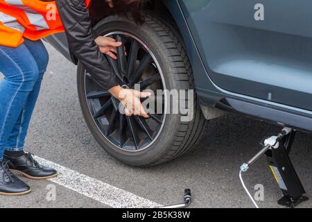 Woman in a orange safety vest is changing a wheel in broken car on the road. Close up female hands to the wheel of a vehicle, trying to change a flat Stock Photo