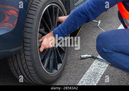 A man in a orange safety vest changes a flat tire on a road. Closeup mans hands to the wheel of a broken car. Replacement of a wheel using skrewdriver Stock Photo