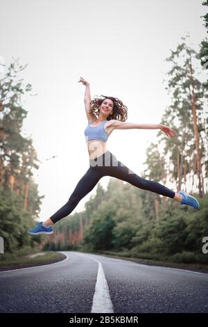 young beautiful brunette girl jumping in twine with background  asphalt road and pine forest Sportive woman showing happiness moments. Dancer training Stock Photo