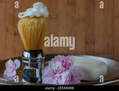 Shaving brush with foarm on top and geranium flowers of tender pink color and white soap on the side on aged steel vase on wooden background. Skin car Stock Photo