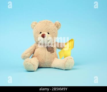 Cute brown teddy bear with a patch holds a silk yellow ribbon in the shape of a loop on a blue background, concept of the fight against childhood canc Stock Photo