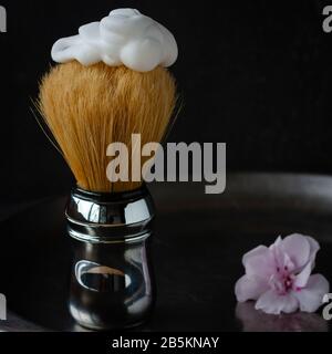 Shaving brush with foarm on top and geranium flowers of tender pink color on the side on aged steel vase on black background. Skin care for men. Chiar Stock Photo
