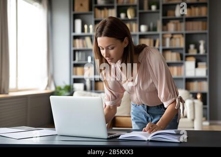 Pleasant young woman working on laptop, standing at table. Stock Photo