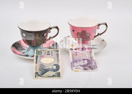 East meets west travel concept with decorated tea coffee cups with 20 british pound note and 1000 yen note on saucers Stock Photo
