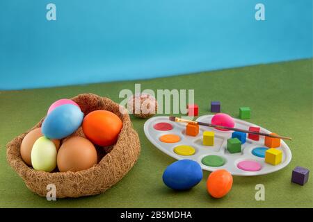 Colored easter eggs in the nest of straw. Palette with a brush for painting Easter eggs. Multi-colored cubes around. Green grass carpet and blue backg Stock Photo