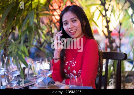 A young Vietnamese woman wearing a traditional red Ao Dai dress using her mobile phone Stock Photo
