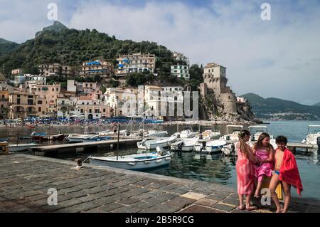 Summertime at the waterfront in Citara, the Amalfi Coast. Three young kids on the jetty. Boats, the beach, and buildings rising high on a blue-sky day Stock Photo