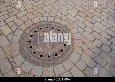 A cast iron manhole cover with a Prague coat of arms and the bridge tower depicted on the round plate , Charles Bridge - Karlův most, Prague. Stock Photo