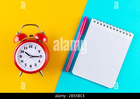 Retro alarm clock next to a pen and notebook on a blue and yellow background. Studying at school and university. Time management concept, business pla Stock Photo